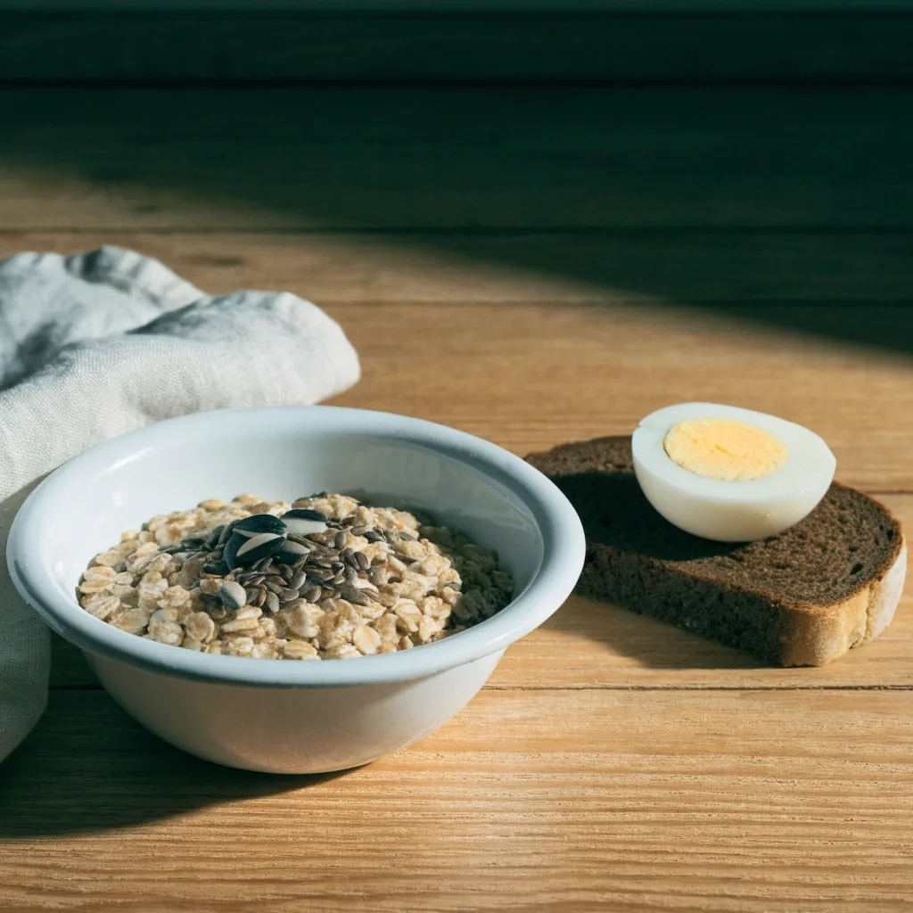 A shallow enamel bowl of oats with seeds next to a slice of rye bread with egg, shot flat-lay on bare oak in low morning light